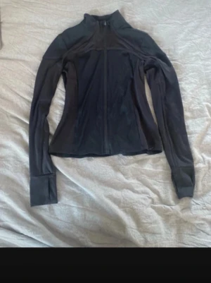 Svart träningsjacka med dragkedja - Säljer en svart träningsjacka med hel dragkedja och hög krage. Jackan har långa ärmar med tumhål och ribbade detaljer på axlar och överdel. Perfekt för löpning eller gym. Materialet är stretchigt och följsamt.