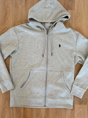 Grå hoodie från Polo Ralph Lauren - Snygg grå zip-hoodie från Polo Ralph Lauren med klassisk huva och dragsko. Broderad logga på bröstet och två stora fickor framtill. Perfekt för en chill och stilren look. Mjuk bomullsblandning som känns skön mot huden.