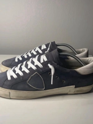 Philippe Model blå låga sneakers - Snygga blå sneakers från Philippe Model med vita detaljer och vita skosnören. Skorna har en låg siluett, vit sula och är tillverkade i mocka och läder. Klassisk design med logotypdetalj på sidan och bekväm passform.