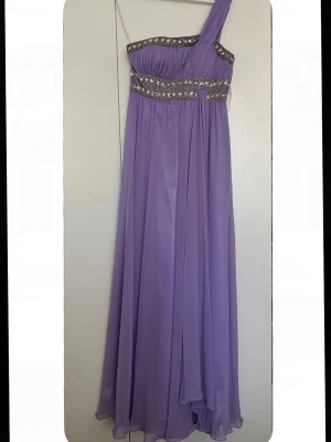 Lila långklänning med paljettdetaljer - Snygg lila långklänning med oneshoulder och glittriga paljettdetaljer vid midjan och över bysten. Klänningen har ett svepande, luftigt tyg som ger en elegant look. Perfekt för bal eller festliga tillfällen.