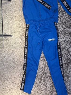 Blå mjukisbyxor från Hoodrich - Snygga blå mjukisbyxor från Hoodrich med svarta sidor och vit logga längs benen. Byxorna har dragsko i midjan och smal passform. Perfekta för en chill och streetwear-inspirerad look.