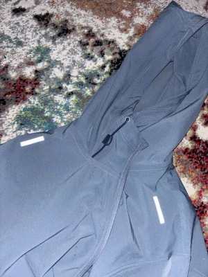 Grå vindjacka från Under Armour - Snygg grå vindjacka från Under Armour med huva och dragkedja. Jackan har reflekterande detaljer på axlarna och en praktisk ficka med dragkedja på bröstet. Tillverkad för den perfekta vädret inför sommaren/våren. Använt max 1, 2 gånger eftersom den var inte den perfekta passformen. Köpte för 1200kr. Priset kan diskuteras ner.