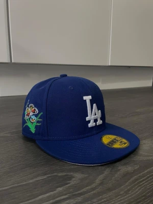 Blå LA keps med fjärilar från New Era - Säljer en blå snapback keps från New Era med LA-logga framtill i vitt och broderade fjärilar och blommor på sidorna. Klassisk rak skärm och coola detaljer som sticker ut. Perfekt för dig som vill ha en unik och trendig look.