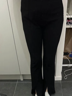 Svarta byxor med slits framtill - Säljer ett par svarta byxor med hög midja och raka ben. Byxorna har en snygg slits framtill vid bensluten som ger en trendig look. Perfekta att styla med sneakers eller klackar. Materialet känns mjukt och stretchigt.