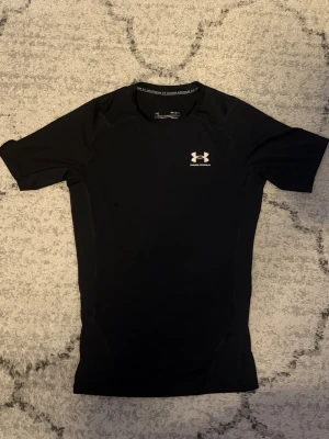 Svart träningströja Under Armour - Säljer en svart, tight och kortärmad träningströja från Under Armour. Tröjan har en diskret logga på bröstet och är tillverkad i ett stretchigt funktionsmaterial som andas. Perfekt för gymmet eller löpning. Är ett litet hål i tröjan.