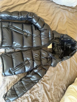 Svart dunjacka från Moncler med huva - Snygg svart dunjacka från Moncler med glansig finish, huva och dragkedjor på fickorna. Jackan har quiltad design och Moncler-logga på ärmen. Perfekt för kalla dagar och riktigt trendig stil. Säljer denna då jag växt ur den och det börjar bli sommar, storlek 1 alltså small men passar mig som är 183 lite kort i ärmarna så skulle tipsa att köpa större storlek om ni är längre än mig! Obs pris kan diskuteras inte hugget i sten:) juste följer även med dustbag (sista bilden) har ingen bild på den!