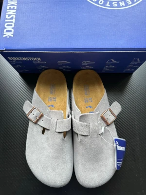 Grå mockatofflor från Birkenstock - Snygga grå tofflor från Birkenstock i mjuk mocka med klassisk design och justerbart spänne i metall. Fotbädden är formad för extra komfort och sulan är i slitstarkt material. Perfekta för dig som vill ha både stil och bekvämlighet.