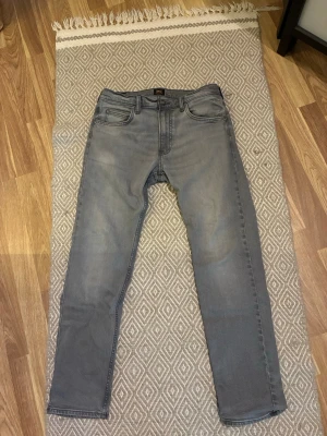 Grå jeans från Lee - Snygga grå jeans från Lee med klassiska fem fickor och svarta detaljer vid midjan. Jeansen har raka ben och är tillverkade i ett mjukt denimtyg som ger en skön känsla. Perfekta för en avslappnad och trendig stil.