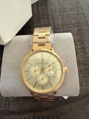 Guldig klocka från Michael Kors - Helt ny Stilren guldig klocka från Michael Kors med metallarmband och rund urtavla. Klockan har tre mindre urtavlor och tydliga guldiga markeringar. Perfekt accessoar för dig som vill ha en lyxig och modern look. All skyddsplast finns kvar