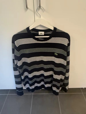 Randig stickad tröja från Lacoste - Snygg stickad tröja från Lacoste i marinblått med vita och gröna ränder. Tröjan har rund halsringning och klassisk Lacoste-logga på bröstet. Perfekt för en stilren och avslappnad look.