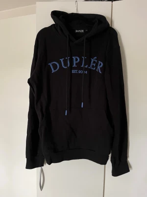 Svart hoodie från Duplér med blå text - Snygg svart hoodie från Duplér med blå text på bröstet och logga på huvan. Klassisk känguruficka framtill, justerbar dragsko i huvan och ribbade muddar. Perfekt för en chill och avslappnad stil.