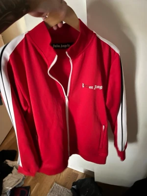 Röd ziptröja från Palm Angels - Palm Angels röd ziptröja med vita och svarta ränder längs ärmarna. Tröjan har hög krage, dragkedja framtill och två fickor. Loggan är tryckt på bröstet. Perfekt för en sportig och trendig look.