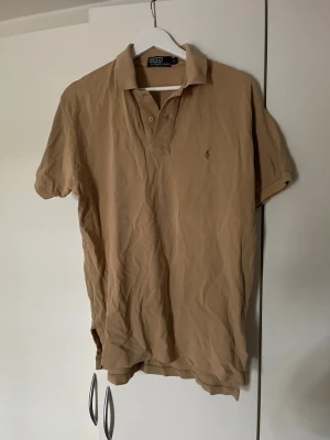 Beige pikétröja från Polo Ralph Lauren - Klassisk beige pikétröja från Polo Ralph Lauren med korta ärmar och krage. Tröjan har knappar vid halsen och den ikoniska lilla broderade loggan på bröstet. Perfekt till en avslappnad men ändå stilren look.