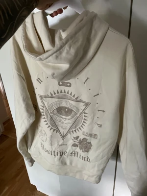 Beige hoodie med tryck och dragkedja - Säljer en beige hoodie  med dragkedja framtill och huva med snören. På ryggen finns ett stort grafiskt tryck med ett öga i en triangel, rosor och texten 'Positive Mind'. Hoodien har kängurufickor och är gjord i mjukt bomullsmaterial.