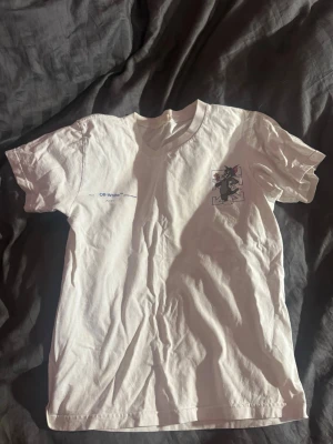 Off-White Tom & Jerry T-shirt vit - Vit T-shirt från Off-White med Tom & Jerry-motiv på ryggen och liten logga framtill. Klassisk passform med rund hals och korta ärmar. Stora blå grafiska detaljer på baksidan och tryckt text.