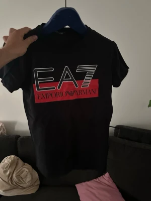 Svart EA7 Emporio Armani t-shirt - Snygg svart t-shirt från EA7 Emporio Armani med stor logga och röd detalj framtill. Klassisk passform och korta ärmar. Perfekt för dig som gillar stilrena märkeskläder.