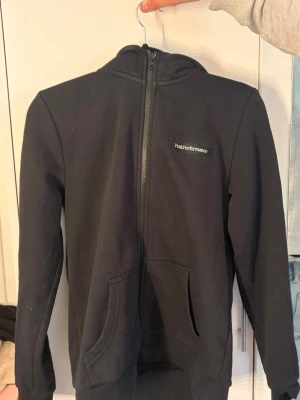 Svart Peak Performance hoodie - Svart hoodie från Peak Performance med dragkedja framtill och fickor på magen. Klassisk design med logga på bröstet och huva. Perfekt för en avslappnad och sportig stil. Mjuk bomullsblandning som känns skön mot huden.