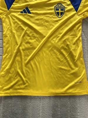 Svensk landslagströja Adidas gul - Gul svensk landslagströja från Adidas med blå detaljer och Sveriges landslagsemblem på bröstet. Tillverkad i lätt och ventilerande ClimaCool-material, perfekt för träning eller match. Klassisk passform med korta ärmar och blå Adidas-logga.