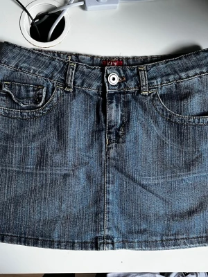 Blå jeanskjol från Fly Girl - Snygg blå jeanskjol från Fly Girl med klassisk femficksdesign, bälteshällor och knappstängning fram. Kjolen är kort och har bakfickor med knappdetaljer. Perfekt för en trendig och avslappnad look. Kjolen är köpt i usa och är i storlek 44 (motsvarar ca 34-36 skulle jag säga)