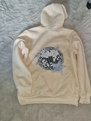 Gul hoodie med discokula och stjärnor - Säljer en gul hoodie med huva och ribbade muddar. På ryggen finns ett stort tryck av en discokula i svart och silver, omgiven av glittriga stjärnor. Hoodien är i mjukt material, perfekt för en cool och trendig look. Skulle säga att den är lite liten i storleken. Aldrig använd eftersom den var för liten.