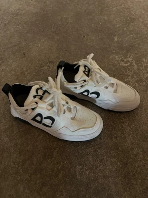 Vita  sneakers med svart detalj - Skorna sitter som 42! Chunky vita sneakers med svart logga på sidan och svarta detaljer vid hälen. Skorna har tjock vit sula, perforerad tå och breda vita skosnören. Tillverkade i syntetmaterial och textil för en sportig look.