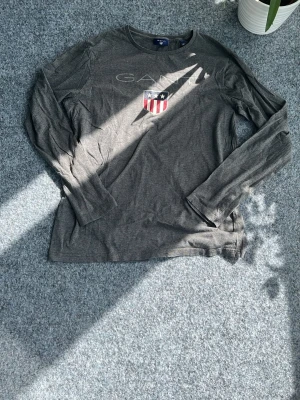 Mörkgrå långärmad tröja från Gant - Snygg mörkgrå långärmad tröja från Gant med rund halsringning. På bröstet finns ett tryckt Gant-logo och ett amerikanskt sköldmärke i rött, vitt och blått. Perfekt till jeans eller chinos för en avslappnad look.