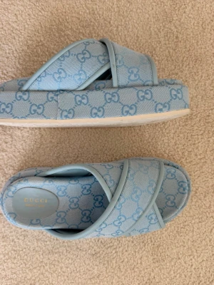Blå sandaler  - Snygga blå sandaler från Gucci med klassiskt GG-logomönster över hela skon. Modellen har korsade remmar och en chunky sula i vitt med präglad logga undertill. Tillverkade i textil och gummi, perfekta för en trendig look.