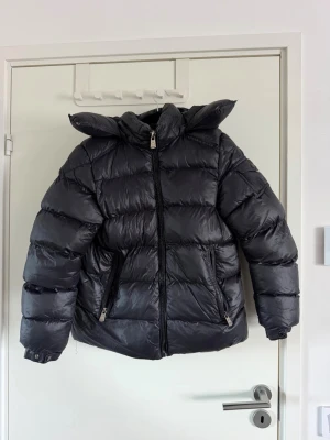 Svart pufferjacka med huva - Säljer en svart pufferjacka med stor huva och dragkedja framtill. Jackan har två snedställda fickor med dragkedja och breda, vadderade paneler för extra värme. Perfekt för kalla vinterdagar och riktigt skön att bära. Loggan finns på ärmen. Använd