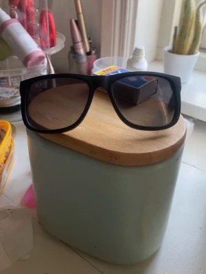 Svarta Ray-Ban solglasögon - Snygga svarta solglasögon från Ray-Ban med bred båge och fyrkantig form. Klassisk logga på sidan och mörka glas. Perfekta för att höja din stil på sommaren eller när solen skiner.