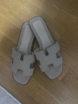 Beige slip-in sandaler med H-rem - Snygga beige slip-in sandaler med breda H-formade remmar och fyrkantig tå. Tillverkade i ett mjukt, strukturerat material som liknar läder. Perfekta för en avslappnad och trendig look till sommaren. jag har köpt dem i Alanya  i Turkiet på en bazar
