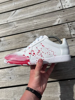 Maison margiela - Unik vit sneaker med handmålad röd färgstänk och penseldrag längs sidan och sulan. Skon har klassisk låg profil, vita snören och sula i gummi. Perfekt för dig som vill sticka ut med en kreativ och personlig stil.