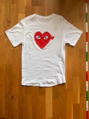 Vit Comme des Garçons Play t-shirt - Vit t-shirt från Comme des Garçons Play med det ikoniska röda hjärtat med ögon tryckt på bröstet. Klassisk passform och rund halsringning. Tillverkad i mjuk bomull och har korta ärmar. Perfekt för en clean och stilren look.