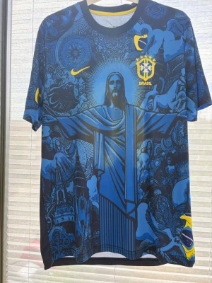 Blå Brasilien fotbollströja Nike - Unik blå fotbollströja från Nike med Brasilien CBF-emblem och detaljerat tryck av Kristusstatyn och brasilianska motiv. Tröjan har gula detaljer och rund hals. Perfekt för dig som vill sticka ut på planen eller heja på landslaget.