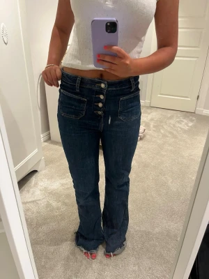 Mörkblå bootcut jeans  - Säljer ett par mörkblå bootcut jeans med hög midja och fyra synliga knappar framtill. Jeansen har stora fickor fram och råa, fransiga benslut för en trendig look. Perfekta till crop tops och sneakers. Materialet är klassisk denim.