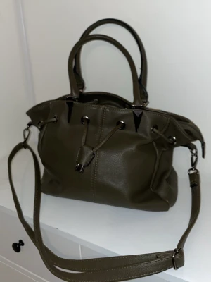 Olivgrön handväska från Eleganci - Vintage olivgrön handväska från Eleganci i konstläder. Väskan har både handtag och en avtagbar axelrem, samt detaljer i silverfärgad metall. Stängs med dragkedja och har dekorativa snörningsdetaljer framtill. 