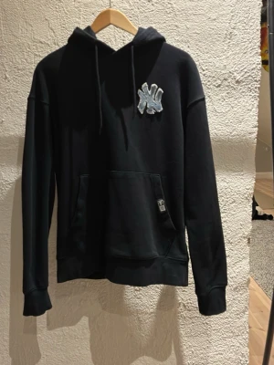 Svart hoodie med NY-logga - Svart hoodie från med stor NY-logga i vitt och blått på bröstet. Klassisk känguruficka framtill och justerbar huva med snören. Tillverkad i mjukt sweatshirtmaterial, perfekt för en avslappnad streetwear-look. Egen gjord NY märke på en vanlig svart hoodie. Använd ungefär 20 gånger. Hör av er vid frågor😄