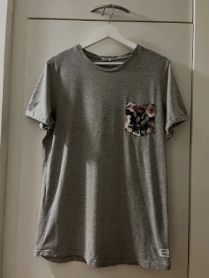 Grå T-shirt från Jack & Jones - Storlek L - Säljer en snygg grå T-shirt från Jack & Jones! Modell: jjorPLAM TEE SS CREW NECK. Kortärmad. Rund hals (O-neck) Praktisk bröstficka med blommigt mönster som snygg detalj! Material: 65 % Bomull och 35 % Polyester. Varumärke: Originals by Jack & Jones. Storlek: L. Säljs i bra skick! Kika gärna in på mina andra annonser, jag säljer även flera klädesplagg! Hör av dig vid frågor eller intresse!