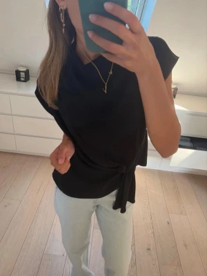Svart omlottopp med knyt - Stilren svart omlottopp med knyt i sidan och korta ärmar. Perfekt till jeans eller kjol för en enkel men snygg look. Tidlös design utan synliga mönster eller loggor.