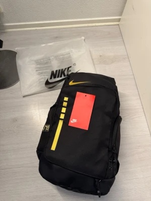 Svart Nike Elite bag ryggsäck - Snygg svart Nike Elite bag ryggsäck med gula detaljer och logga. Ryggsäcken har flera fack, vadderade axelband med silver- och guldfärgade detaljer samt en praktisk topphandtag. Perfekt för skola, träning eller vardag. Tillverkad i slitstarkt material. Allt följer med. Inte använd en ända gång. Finns fler bilder. Fraktar samma dag eller dagen efter :)