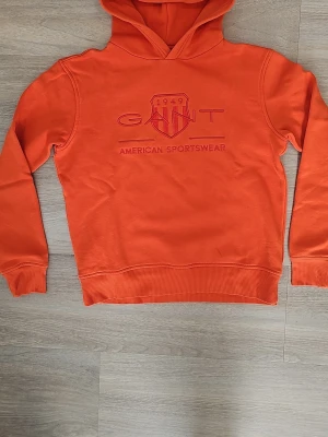 Orange hoodie från GANT - Snygg orange hoodie från GANT med broderad logga och texten 'American Sportswear' på bröstet. Klassisk huva och ribbade muddar vid ärmslut och nederkant. Tillverkad i mjukt bomullsmaterial som känns skönt mot huden. Perfekt för en avslappnad och sportig stil. Storlek 170 för 13-14 åringar. Pris kan diskuteras, hör gärna av er!