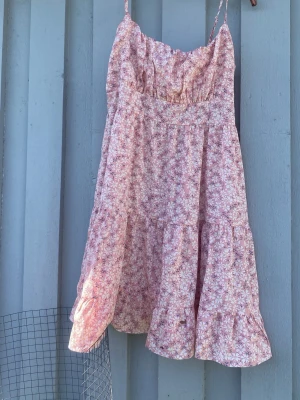 Blommig rosa sommarklänning - Söt kortklänning med smala axelband och volangkant. Klänningen är rosa med små vita blommor och har en smockad rygg för skön passform. Perfekt för varma dagar och har en luftig, flowy känsla.