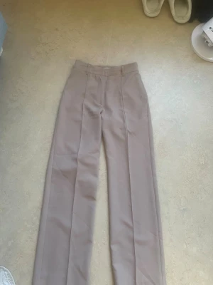Beige vida kostymbyxor - Snygga beige kostymbyxor med hög midja och vida ben. Klassisk pressveck framtill och hällor för skärp. Perfekta för en stilren och trendig look. Materialet ser ut att vara en mjuk och följsam blandning, troligtvis polyester eller viskos. Oanvänd 
