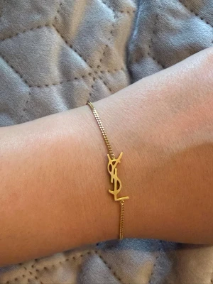 Guldigt YSL armband - Snyggt och stilrent armband i guldton från Yves Saint Laurent med den ikoniska YSL-logotypen som detalj. Tunt kedjearmband med justerbar längd och elegant lås. Perfekt accessoar för dig som gillar lyxiga och minimalistiska smycken.