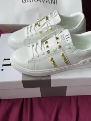 Vita Valentino sneakers med nitar - Snygga vita sneakers från Valentino Garavani med guldfärgade nitar på sidorna och över vristen. Skorna är i läder med klassisk låg profil och har en unik sula med rutmönster. Perfekta för dig som vill sticka ut med exklusiv design.