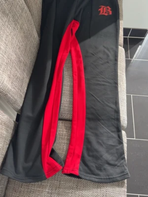 Svarta mjukisbyxor med röd detalj - Säljer ett par svarta mjukisbyxor med röd kontrastpanel längs benen och ett tryckt B-märke vid höften. Byxorna har raka ben och är tillverkade i ett mjukt, stretchigt material som känns skönt mot huden. Perfekta för en avslappnad och sportig stil.