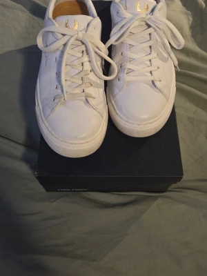Vita Fred Perry sneakers i läder - Snygga vita sneakers från Fred Perry i läder med guldfärgade detaljer och klassisk logga på sidan. Skorna har vita snören och en stilren, låg profil som passar till det mesta. Perfekta för dig som gillar enkel och clean stil.
