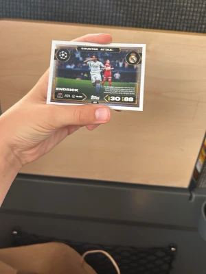 Match Attax Endrick Real Madrid kort - Samlarbild från Match Attax med Endrick i Real Madrid, motivet visar spelaren i vit matchdräkt på fotbollsplan. Kortet har gulddetaljer och texten 'Counter Attack'. Baksidan har Real Madrids logga och Match Attax-design. Perfekt för dig som gillar fotboll och samlarkort.