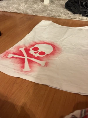 Vit tubtopp med dödskalletryck - Snygg vitt linne med stort rött dödskalle- och korsben-tryck framtill. Toppen har en lite kurvigare passform. Materialet känns som bomull och designen är perfekt för dig som gillar alternativ och edgy stil.