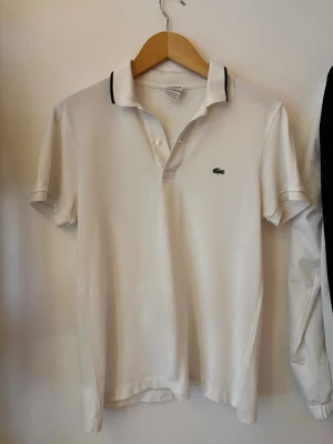 Vit pikétröja från Lacoste - Vit pikétröja från Lacoste med klassisk krage och korta ärmar. Tröjan har svarta detaljer på kragen och den ikoniska gröna krokodilloggan på bröstet. Perfekt för en clean och sportig look.
