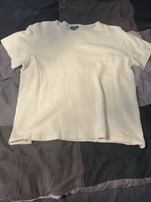 Ljusbeige ribbad t-shirt - En stilren ljusbeige t-shirt med ribbad struktur och rund hals. T-shirten har korta ärmar och en avslappnad passform, perfekt för en enkel och clean look. Materialet känns mjukt och bekvämt mot huden.